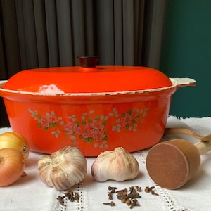 May include: An orange enamel Dutch oven with a black handle and floral design. Onions, garlic, cloves, and a wooden mallet and spoon are arranged on a white tablecloth, suggesting a cooking theme.