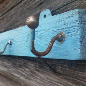 Puede incluir: Un perchero de madera rústico con tres ganchos de metal pintados de azul claro. Los ganchos son de metal y tienen una parte superior redondeada.