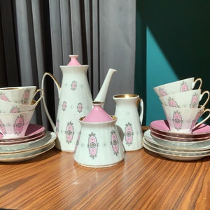 K&ouml;nnte beinhalten: Ein Vintage-Teeservice mit wei&szlig;en Porzellanst&uuml;cken mit rosa und grauen dekorativen Mustern. Das Set umfasst Tassen, Untertassen, eine Teekanne, einen Milchgie&szlig;er und eine Zuckerdose. Goldene Verzierungen akzentuieren die R&auml;nder der Tassen und Untertassen.