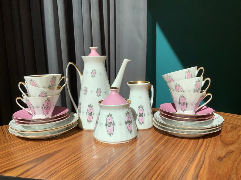 K&ouml;nnte beinhalten: Ein Vintage-Teeservice mit wei&szlig;en Porzellanst&uuml;cken mit rosa und grauen dekorativen Mustern. Das Set umfasst Tassen, Untertassen, eine Teekanne, einen Milchgie&szlig;er und eine Zuckerdose. Goldene Verzierungen akzentuieren die R&auml;nder der Tassen und Untertassen.