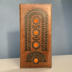 Könnte beinhalten: Eine rechteckige, kupferfarbene Geldbörse mit geprägtem geometrischen Design und orangefarbenen Akzenten. Die Geldbörse hat einen dekorativen Rand und ein zentrales Muster aus Bögen, Kreisen und sternförmigen Formen. Das Design ist symmetrisch und detailliert.