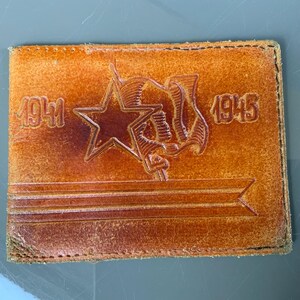 May include: A rectangular, tan leather wallet with embossed imagery. The design includes a star, a flag, and the years "1941" and "1945". The edges of the wallet are stitched, and the leather has a worn appearance.