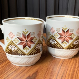 Könnte beinhalten: Zwei weiße Keramikbecher mit goldfarbenem Rand und einem geometrischen Muster in Braun, Rosa und Gold. Die Becher stehen auf einer Holzoberfläche.