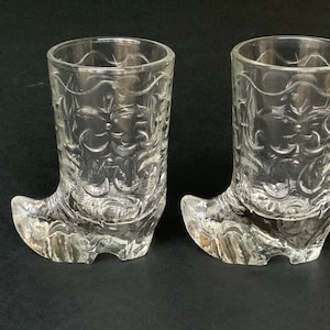 May include: Two clear glass shot glasses shaped like cowboy boots. The boots have a detailed floral pattern etched into the glass. The shot glasses are displayed on a dark surface.