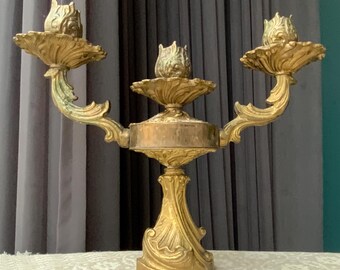 Könnte beinhalten: Ein goldfarbener Metall-Kandelaber mit drei Kerzenhaltern. Der Kandelaber hat ein dekoratives, verziertes Design mit einem runden Sockel und drei Armen, die sich nach oben biegen.