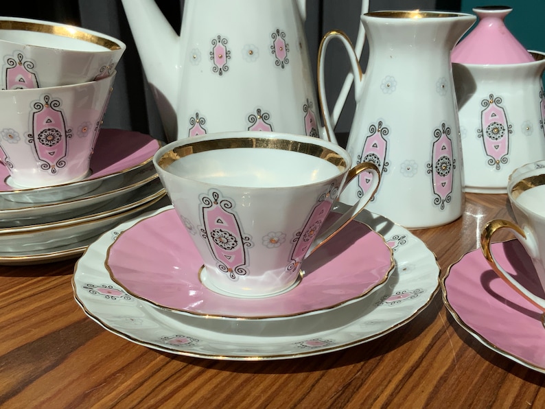 K&ouml;nnte beinhalten: Vintage Teeservice mit wei&szlig;en Porzellantassen, Untertassen und Servierteilen. Das Set hat ein rosa-schwarzes Dekor mit goldenen Verzierungen. Enth&auml;lt Tassen, Untertassen, eine Teekanne, einen Sahnek&auml;nnchen und eine Zuckerdose.