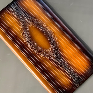 Puede incluir: Un tablero de backgammon rectangular de madera con un acabado de dos tonos. El centro presenta un diseño ovalado, ornamentado y en relieve. El tablero tiene un patrón de rayas y un borde marrón oscuro.