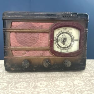 Peut inclure: Une radio ancienne avec un boîtier marron foncé et noir. La radio a un couvercle de haut-parleur en tissu rouge et un cadran circulaire avec un graphique d'une personne à cheval. Trois boutons sont situés en bas de la radio.