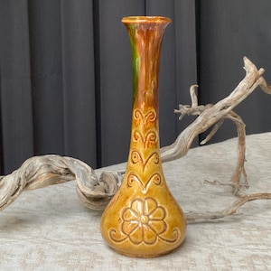 May include: A tall, slender ceramic bud vase with a glossy mustard yellow and brown glaze. The vase features a relief-molded floral design at the base and a series of repeating scroll patterns extending upward along the neck toward the flared rim.