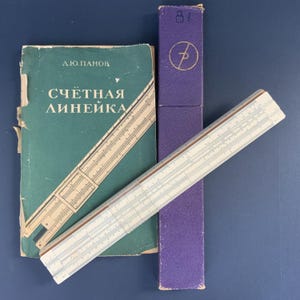Puede incluir: Una regla de cálculo vintage en una caja púrpura con un libro titulado "Счётная Линейка" de Д.Ю. Панов. La regla de cálculo está hecha de plástico blanco con marcas negras. El libro es verde con letras blancas.