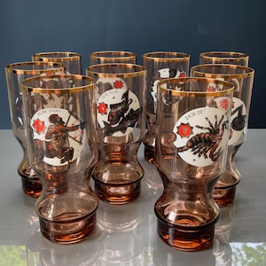 May include: A set of ten vintage brown-tinted drinking glasses with gold rims. Each glass features a different zodiac sign illustration in white and red. The glasses are arranged on a reflective surface against a dark blue background.