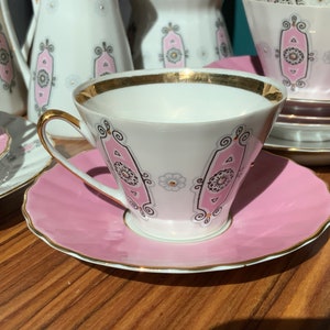 K&ouml;nnte beinhalten: Ein wei&szlig;es Teeservice mit rosa und goldenen Verzierungen. Die Tassen haben goldene R&auml;nder und Henkel, die Untertassen sind rosa. Weitere Teile des Sets, wie eine Teekanne, sind ebenfalls zu sehen.