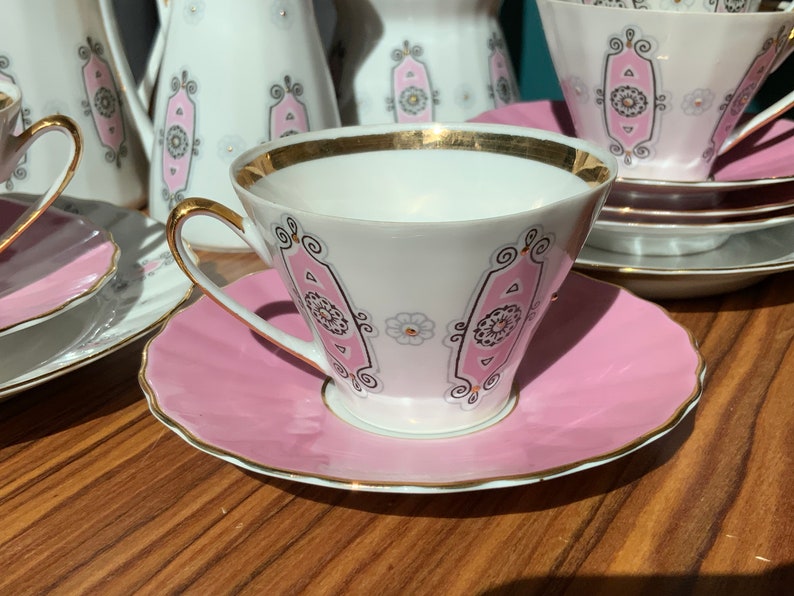K&ouml;nnte beinhalten: Ein wei&szlig;es Teeservice mit rosa und goldenen Verzierungen. Die Tassen haben goldene R&auml;nder und Henkel, die Untertassen sind rosa. Weitere Teile des Sets, wie eine Teekanne, sind ebenfalls zu sehen.
