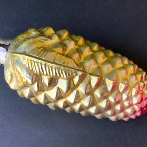 Könnte beinhalten: Ein goldener gläserner Weihnachtsbaumschmuck in Form einer Tannenzapfen mit roter Spitze. Der Schmuck hat eine strukturierte Oberfläche und ein goldenes Blattdesign.