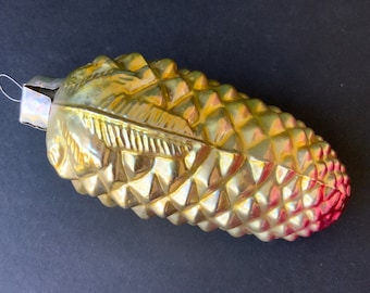 May include: A gold glass Christmas ornament shaped like a pine cone with a red tip. The ornament has a textured surface and a gold leaf design.