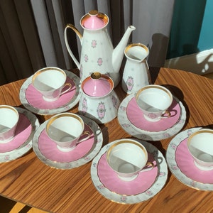 K&ouml;nnte beinhalten: Vintage Teeservice mit wei&szlig;em und rosa Blumenmuster. Das Set enth&auml;lt eine Teekanne, einen Sahnek&auml;nnchen, eine Zuckerdose und sechs Teetassen mit Untertassen. Jedes St&uuml;ck hat einen goldenen Rand. Das Set ist auf einem Holztisch angeordnet.