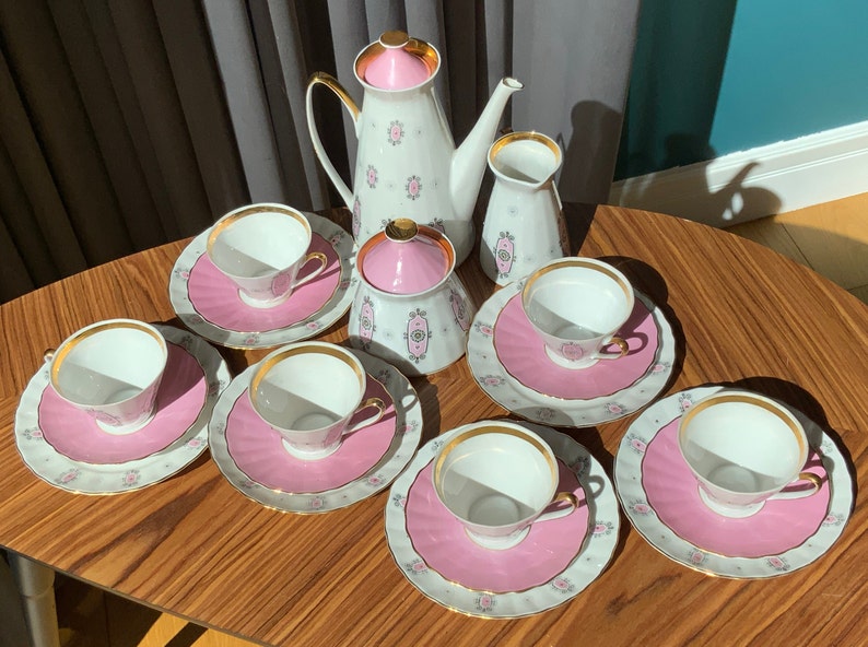 K&ouml;nnte beinhalten: Vintage Teeservice mit wei&szlig;em und rosa Blumenmuster. Das Set enth&auml;lt eine Teekanne, einen Sahnek&auml;nnchen, eine Zuckerdose und sechs Teetassen mit Untertassen. Jedes St&uuml;ck hat einen goldenen Rand. Das Set ist auf einem Holztisch angeordnet.