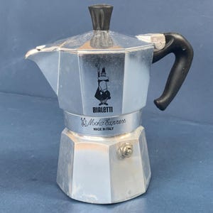 Pode incluir: Uma cafeteira italiana Bialetti Moka Express prateada com uma pega e botão pretos. O logótipo Bialetti, as palavras "Moka Express" e "Made in Italy" são visíveis. Ideal para preparar café expresso.