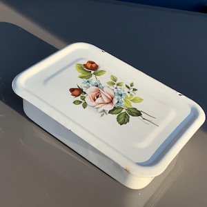 May include: White rectangular metal container with a lid. The lid features a floral design with a large pink rose, smaller orange buds, and blue and white accent flowers. Green leaves complete the arrangement.