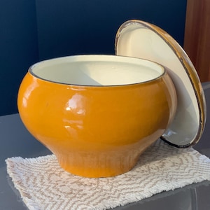 Könnte beinhalten: Ein runder, orangefarbener Emailletopf mit passendem Deckel. Der Topf hat einen weißen Innenraum und steht auf einem beigefarbenen, gemusterten Platzdeckchen. Der Deckel liegt daneben und gibt den Blick ins Innere frei.