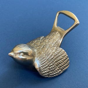 Può includere: Un apribottiglie in metallo argentato a forma di uccello con ali dettagliate. L'apribottiglie ha un design classico con un anello in alto.