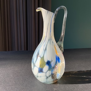 Op de afbeelding: Een witte glazen kan met een heldere handvat. De kan heeft een parelmoerachtige afwerking met blauwe, gele en gouden swirls en vlekken.
