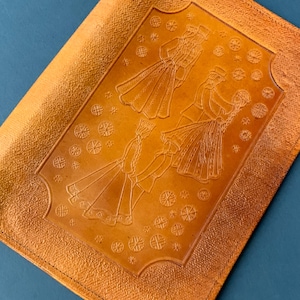May include: A golden-brown leather-like item with an embossed design of figures in traditional clothing. The central design features three pairs of figures, possibly dancing, surrounded by decorative circular patterns. The item has a textured surface.