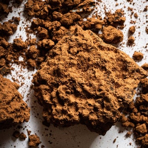 Puede incluir: Primer plano de un mont&oacute;n de polvo de cacao marr&oacute;n sobre una superficie blanca. El polvo es de varios tama&ntilde;os y formas, algunos est&aacute;n agrupados.
