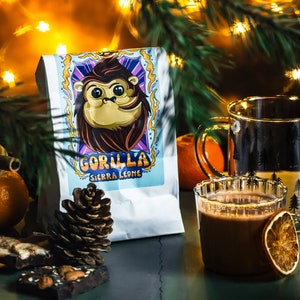 Puede incluir: Una bolsa blanca de granos de caf&eacute; con un gorila de dibujos animados en la parte delantera. La bolsa dice "Gorilla Sierra Leone". Hay una taza de chocolate caliente con una rodaja de naranja, una pi&ntilde;a y un poco de corteza de chocolate sobre una mesa verde.