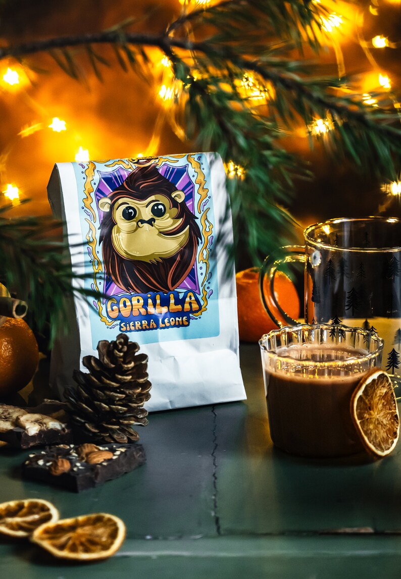 Puede incluir: Una bolsa blanca de granos de caf&eacute; con un gorila de dibujos animados en la parte delantera. La bolsa dice "Gorilla Sierra Leone". Hay una taza de chocolate caliente con una rodaja de naranja, una pi&ntilde;a y un poco de corteza de chocolate sobre una mesa verde.