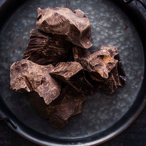 Puede incluir: Primer plano de trozos de chocolate marr&oacute;n oscuro en una bandeja de metal. El chocolate est&aacute; roto en trozos irregulares, revelando su rico color oscuro y textura.