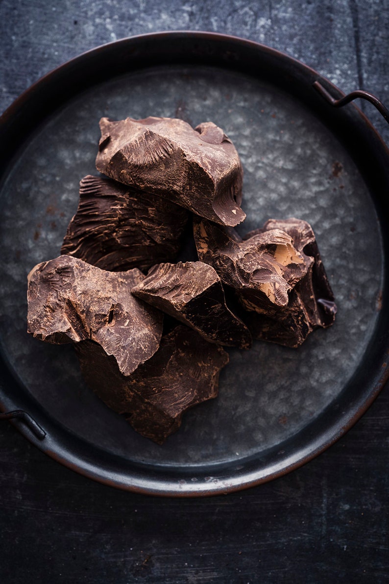 Puede incluir: Primer plano de trozos de chocolate marr&oacute;n oscuro en una bandeja de metal. El chocolate est&aacute; roto en trozos irregulares, revelando su rico color oscuro y textura.