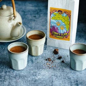 May include: Three white ceramic mugs filled with a dark brown liquid, likely hot chocolate, on a blue textured surface. A white paper bag with the text "Lion's Claw Congo" is in the background. There are also pieces of broken dark chocolate on the surface.