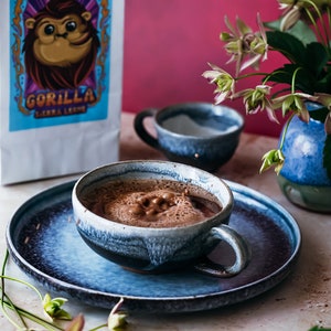 Puede incluir: Una taza de cer&aacute;mica azul llena de chocolate caliente espumoso sobre un platillo de cer&aacute;mica azul. La taza tiene un esmalte azul claro con vetas azules m&aacute;s oscuras. El platillo tiene un esmalte similar. Una bolsa de granos de caf&eacute; con un logotipo de gorila est&aacute; en el fondo.