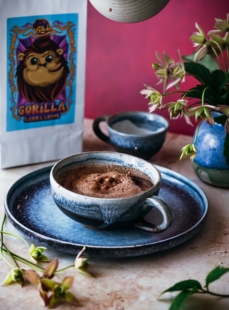 Puede incluir: Una taza de cer&aacute;mica azul llena de chocolate caliente espumoso sobre un platillo de cer&aacute;mica azul. La taza tiene un esmalte azul claro con vetas azules m&aacute;s oscuras. El platillo tiene un esmalte similar. Una bolsa de granos de caf&eacute; con un logotipo de gorila est&aacute; en el fondo.