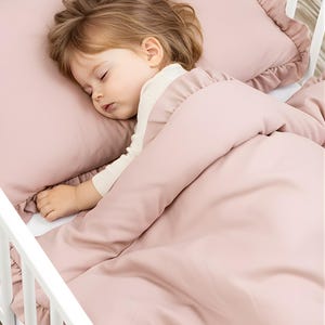 Może przedstawiać: Śpiące dziecko w białym łóżeczku, otulone miękką, różową kołdrą i poduszką. Poduszka i kołdra mają marszczone krawędzie. Dziecko ma na sobie białą koszulkę z długim rękawem.