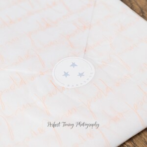 May include: White tissue paper with a pink pattern of the words "Baby Hugs" repeated. A white circle sticker with three blue stars and the text "Baby Hugs" is on the tissue paper.