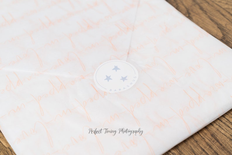 May include: White tissue paper with a pink pattern of the words "Baby Hugs" repeated. A white circle sticker with three blue stars and the text "Baby Hugs" is on the tissue paper.