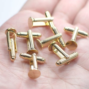 May include: A collection of gold-colored cufflinks. The cufflinks have a classic design with a flat, circular face and a straight bar connecting to a post. The metal has a shiny finish. The cufflinks are scattered on a light background.