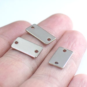 May include: Three silver rectangular metal tags with two holes each. The tags are on a hand.