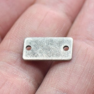 Puede incluir: Un pequeño dije rectangular de metal con un acabado plateado envejecido. El dije tiene esquinas redondeadas y dos pequeños agujeros, uno en cada extremo. La superficie muestra una textura sutil y desgaste, lo que sugiere una estética antigua o vintage. Ideal para la fabricación de joyas o manualidades.
