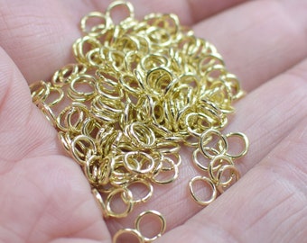 Puede incluir: Un montón de anillas de salto doradas, un tipo de hallazgo de joyería, en una mano. Las anillas son pequeñas y delicadas, y son perfectas para hacer joyas.