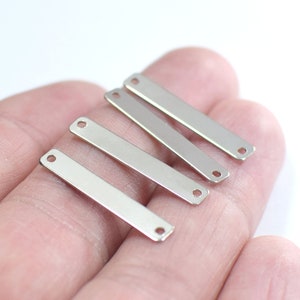 Könnte beinhalten: Vier silberne Metall-Bar-Anhänger mit kleinen Löchern an jedem Ende. Die Anhänger sind in einem Stapel auf einer Hand angeordnet.