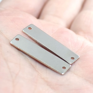 Pode incluir: Dois pingentes retangulares de metal prateado com um furo em cada extremidade. Os pingentes estão sobre uma mão.