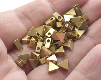 May include: A pile of gold-colored metal triangle-shaped beads with a hole in the center. The beads are scattered on a hand.