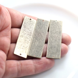 Pode incluir: Três etiquetas retangulares de metal com um acabamento texturizado e envelhecido. Cada etiqueta tem um furo na parte superior e inferior. As etiquetas são seguradas em uma mão contra um fundo branco. O metal tem uma cor dourada antiga.