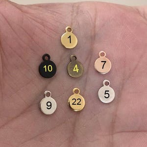 Könnte beinhalten: Eine Sammlung kleiner, runder Metallanhänger in verschiedenen Farben, darunter Gold, Silber und Schwarz. Jeder Anhänger hat eine Nummer von 1 bis 22 und eine kleine Schlaufe zum Befestigen an einer Kette oder einem Armband. Die Anhänger werden auf einer Hand präsentiert.