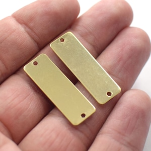May include: Two rectangular, gold-colored metal tags with rounded corners and two small holes. The tags are smooth and shiny, and appear to be new. The tags are held in a person's hand.