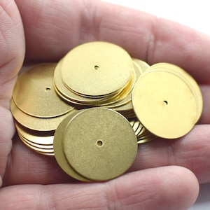 May include: A pile of round, gold-colored metal discs with a small hole in the center. The discs are stacked on top of each other and are being held in a hand.