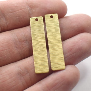 Puede incluir: Dos colgantes de barra de metal de color dorado con una superficie texturizada y un agujero en la parte superior.